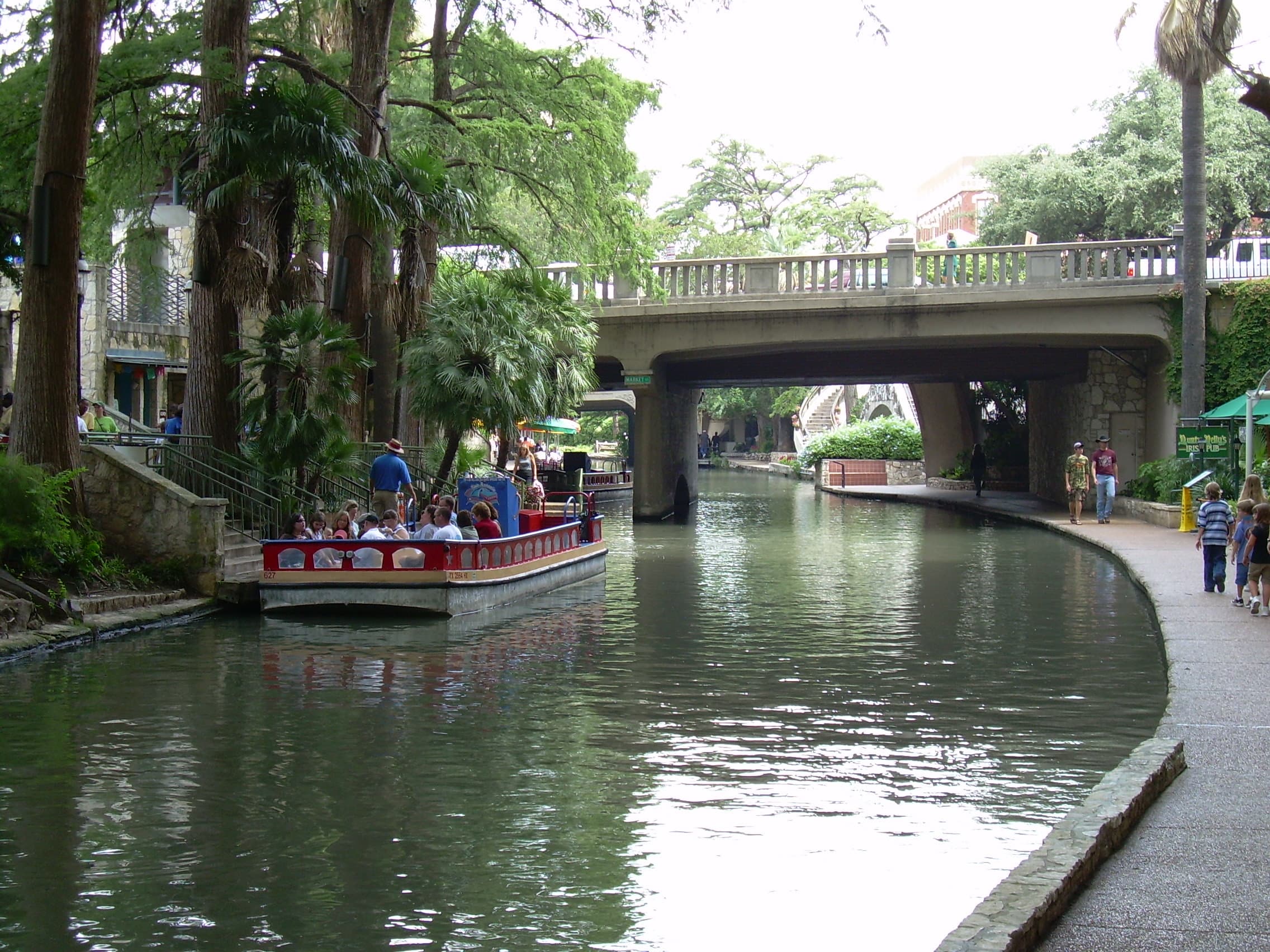 The San Antonio River Walk — pedestrians along the stone walkway beside the river, with arched bridges and lush bald cypress trees overhead.