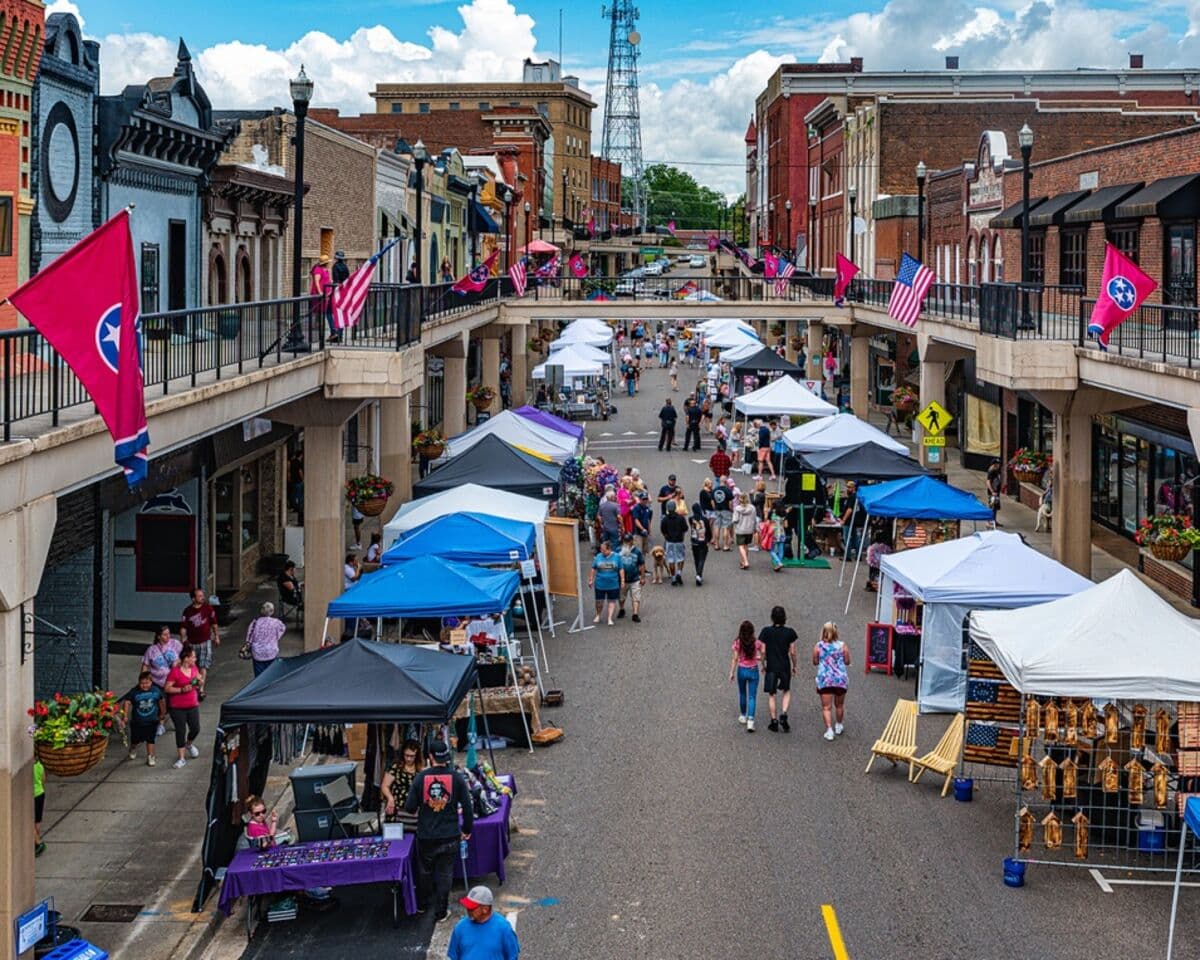 A street festival in downtown Morristown — vendor tents in blue and white running down Main Street under the elevated SkyMart walkways, crowds browsing and walking.
